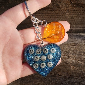 Puede incluir: Un llavero en forma de corazón azul con purpurina con las palabras "DABS ALL DAY" en letras plateadas. El llavero también tiene un amuleto redondo naranja.