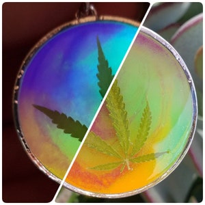 May include: A round, iridescent pendant with a silver rim, featuring a pressed green cannabis leaf. The pendant has a rainbow-like effect with shades of blue, green, and yellow.