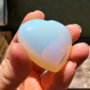 Puede incluir: Un cristal de ópalo iridiscente en forma de corazón, de color blanco lechoso y azul pálido. El cristal es liso y pulido.