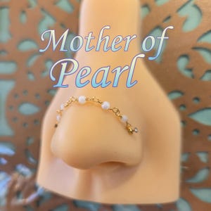 May include: A gold-colored nose ring with small, white pearls. The nose ring is attached to a nose model. The text "Mother of Pearl" is displayed in a cursive font above the nose ring.