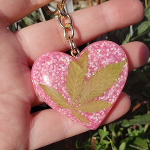 May include: Pink heart-shaped keychain with a pressed leaf inside and glitter embedded in the resin. The keychain is attached to a gold chain.