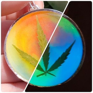May include: A silver pendant with a clear resin dome featuring a green cannabis leaf. The resin changes colour from yellow to blue depending on the light.