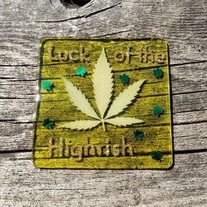 May include: A yellow square coaster with gold glitter and the text "Luck of the Highrish" with a green leaf design in the center.