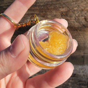 May include: A small, clear glass jar with a gold lid containing a golden yellow substance. The jar is attached to a gold chain.