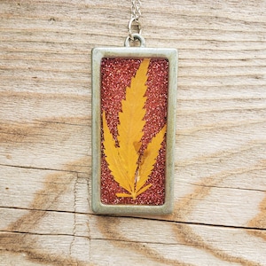 May include: A silver necklace with a rectangular pendant featuring a pressed, dried yellow leaf on a red glitter background.