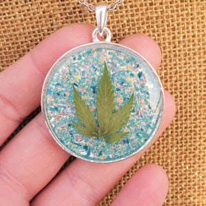 Puede incluir: Un collar de plata con un colgante redondo que presenta una hoja verde prensada encapsulada en resina de purpurina iridiscente.