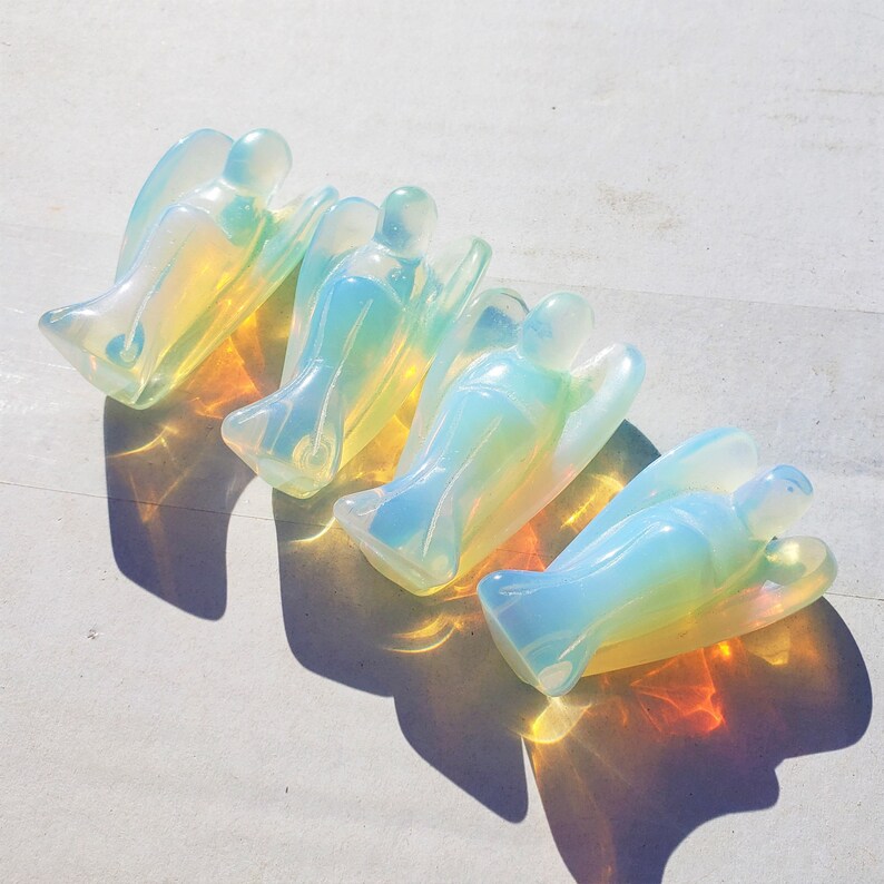May include: Four iridescent opal angel figurines, each with a different shade of blue and yellow, arranged in a row on a white surface.