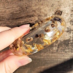 May include: A clear resin bangle bracelet with gold leaf and brown flecks embedded inside.