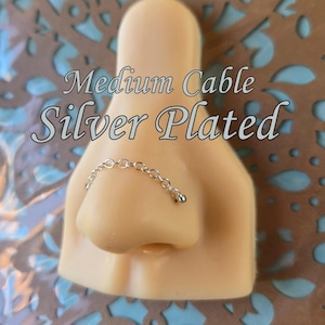 May include: A silver-plated nose chain with a medium cable design. The chain is draped across a nose, with a small bead at the end. The text "Medium Cable Silver Plated" is above the nose chain.