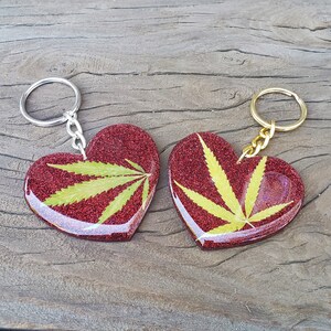 May include: Two red glitter heart-shaped keychains with gold and silver chains. Each heart features a pressed green leaf inside.