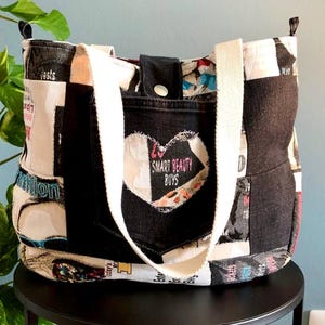 Può includere: Una borsa tote patchwork con un cinturino bianco e una tasca in denim nera. La tasca presenta un design a cuore con la scritta "SMART BEAUTY BOYS". La borsa è realizzata con vari pezzi di tessuto con testo e immagini stampate.