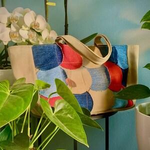 Può includere: Una borsa tote beige con un design patchwork di cerchi in denim, rossi e beige. La borsa ha un manico beige. Piante verdi e orchidee bianche circondano la borsa.