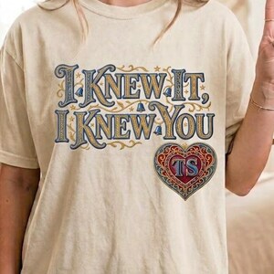May include: An ivory short-sleeve t-shirt featuring the text "I KNEW IT, I KNEW YOU" in a decorative blue and gold font. Below the text is a red, blue, and gold heart-shaped emblem containing the letters "TS" in a stylized, ornate design.