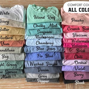May include: A collection of folded t-shirts in various colors labeled with text including White, Ivory, Butter, Mustard, Yam, Sandstone, Bay, Light Green, Blue Spruce, Moss, Island Reef, Chalky Mint, Seafoam, Hydrangea, Chambray, Blue Jean, Flo Blue, Washed Denim, Denim, Gray, Pepper, Peachy, Crunch Berry, Blossom, Watermelon, Heliconia, Brick, Chili, Orchid, Violet, Berry, and Black.