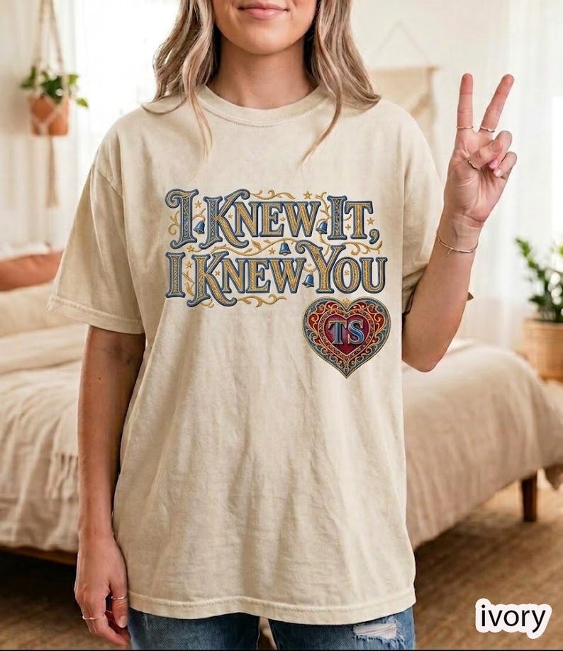 May include: An ivory short-sleeve t-shirt featuring the text "I KNEW IT, I KNEW YOU" in a decorative blue and gold font. Below the text is a red, blue, and gold heart-shaped emblem containing the letters "TS" in a stylized, ornate design.