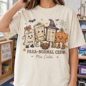 May include: A cream-colored t-shirt featuring a graphic of four cartoon characters: a ghost labeled PARAPRO, a mummy holding a checklist, a clipboard wearing a witch hat, and a star-shaped pumpkin. Text reads PARA-NORMAL CREW and Mrs. Carter.