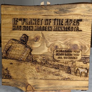 May include: Wooden plaque with carved text and imagery. The text reads "IF 'PLANET OF THE APES' HAD BEEN MADE IN MINNESOTA..." and "GOSH DARN YOU GOSH DARN YOU ALL TO HECK!". The plaque features a carved figure and landscape.