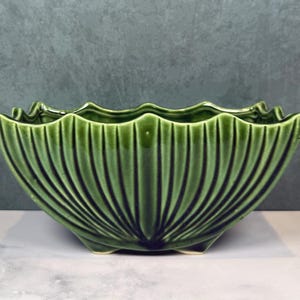 May include: A rectangular ceramic planter with a glossy dark green glaze, featuring a vertical ribbed pattern that radiates from the base. The top edge has a scalloped, wavy design, and the vessel is supported by small, integrated feet at the bottom.