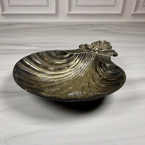 May include: A silver-toned, seashell-shaped dish with a detailed, ridged design. The dish has a decorative, ornate handle. The seashell dish is likely made of metal and is suitable for holding small items.