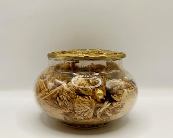 May include: A clear glass jar with a decorative gold lid. The jar is filled with dried flowers and small gold ornaments. The lid has an ornate, embossed design. The jar is sitting on a white surface.