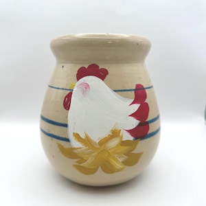 以下が含まれることがあります： 手描きの鶏のデザインが施されたベージュ色のセラミックジャー。鶏は白で、赤いアクセントと黄色のベースがあります。青いストライプがジャーを囲み、丸みを帯びた形をしています。