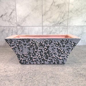 May include: A square ceramic planter with a textured gray and black exterior and a pink interior. The planter has a trapezoidal shape, with the top wider than the base. The background is a light gray tiled wall.