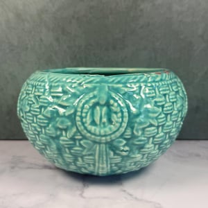 May include: A round, turquoise ceramic bowl featuring a raised, textured basket-weave pattern across its exterior surface. The bowl includes a prominent circular medallion design on the front, with a glossy finish and a wide, open top opening.