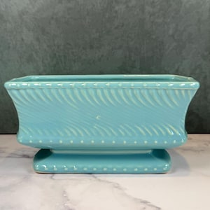 May include: A rectangular turquoise ceramic planter featuring a raised diagonal ribbed texture on the sides, a beaded border along the top rim, and a matching beaded base. The vessel has a flared opening and sits on a pedestal-style foundation.