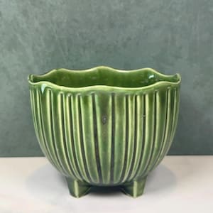 Op de afbeelding: Een kleine, ronde groene keramische plantenpot met een glanzende afwerking en een verticale geribbelde textuur. De pot heeft een golvende rand en staat op drie korte, geïntegreerde voetjes, met een diepe bosgroene kleur en donkere accenten.