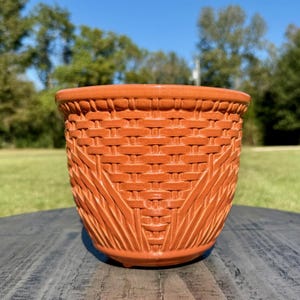 May include: A terracotta-colored ceramic planter with a woven basket design. The planter has a wide, rounded shape and a textured surface, suitable for indoor or outdoor use. The background features a blurred view of green trees and a blue sky.