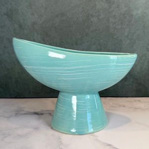 May include: A turquoise ceramic bowl with a pedestal base, featuring a subtle white horizontal streaked pattern across the exterior surface. The bowl has an asymmetrical, elongated oval rim that slopes downward from one side to the other.