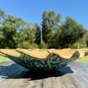 Puede incluir: Un cuenco de cerámica hecho a mano con una forma orgánica única. El cuenco presenta un interior verde moteado y un borde marrón contrastante. El cuenco se exhibe sobre una superficie de madera, con un telón de fondo de árboles verdes y cielo azul.