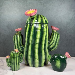 May include: A ceramic cactus-shaped jar with a lid and two smaller cactus-shaped figurines. The large jar features a tall, ribbed green body with a pink flower on top and two side arms, each topped with a pink flower. Two smaller cacti accompany it.