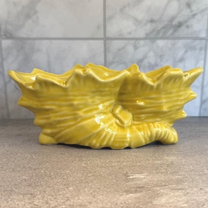 May include: A yellow ceramic seashell-shaped planter. The planter has a textured surface with ridges and a glossy finish. The design is a detailed replica of a seashell, suitable for small plants or decorative use.
