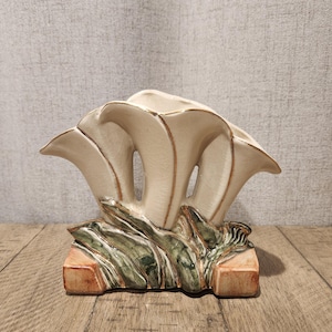 Può includere: Un vaso in ceramica color crema con un design floreale unico. Il vaso presenta molteplici strutture aperte a forma di petalo con bordi marroni, poggianti su una base con accenti verdi e marroni. Un oggetto decorativo per la casa.