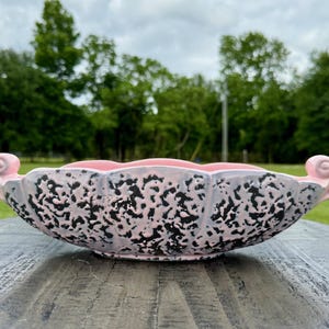 May include: A decorative ceramic bowl with a pink interior and exterior, featuring a speckled black and grey pattern. The bowl has a scalloped edge and two pink, curled handles. The bowl is on a grey wooden surface.