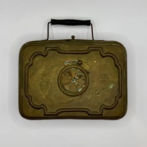 Puede incluir: Antigua fiambrera de metal color latón con asa negra. La caja rectangular tiene un borde decorativo y un diseño circular en la parte delantera. El asa está sujeta a la parte superior con soportes metálicos.