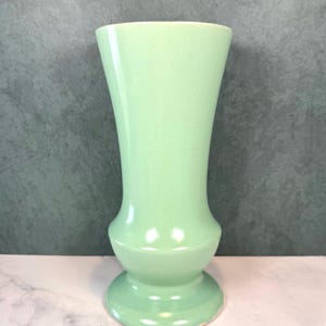 May include: A tall, light mint green ceramic vase with a smooth, glossy finish. The vase features a wide, flared opening at the top, a slender neck that tapers inward, and a rounded, bulbous midsection that rests on a circular, pedestal-style base.