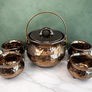Puede incluir: Un conjunto de cinco piezas de cerámica con un acabado martillado en bronce metálico. La colección incluye una olla grande con tapa y una fina asa de metal dorado, además de cuatro cuencos a juego con pequeñas patas y asas laterales.