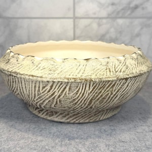 May include: A round ceramic bowl featuring a textured exterior with a cream and dark brown wood grain pattern. The bowl has a scalloped rim and a smooth, solid cream-colored interior. The base is slightly tapered, providing a stable foundation.