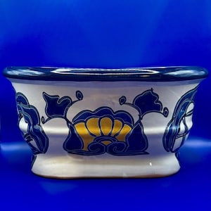 Könnte beinhalten: Ein weißer Keramik-Blumentopf mit blauem und goldenem Blumendesign. Der Pflanztopf hat eine rechteckige Form mit gebogenem Rand und ist mit stilisierten Blumen und Blättern verziert. Der Hintergrund ist einfarbig blau.