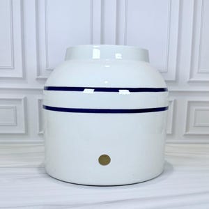 Peut inclure: Un pot en céramique blanc avec deux rayures horizontales bleu marine. Le pot a une forme arrondie avec un sommet légèrement plus étroit et une petite ouverture ronde. Un petit trou doré est visible près du fond.