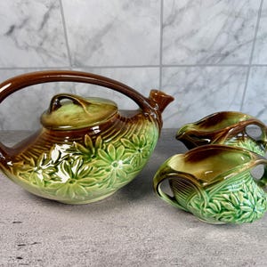 Può includere: Un servizio da tè in ceramica vintage con una teiera con un manico e coperchio grandi e due lattine. Il set è smaltato in tonalità marroni e verdi, con un motivo floreale in rilievo. La teiera ha un beccuccio curvo.