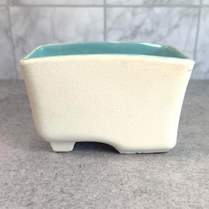 Può includere: Un vaso rettangolare in ceramica con una finitura esterna testurizzata color bianco sporco e uno smalto interno liscio color turchese brillante. Il contenitore presenta quattro piccoli piedini integrati alla base e un'apertura ampia.