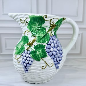 May include: White ceramic pitcher with a woven basket texture. The pitcher is adorned with green grape leaves and clusters of blue grapes. The handle and spout are smooth white. A decorative piece for serving beverages.