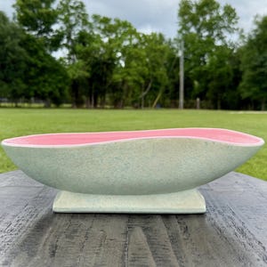 Puede incluir: Una jardinera de cerámica de mediados de siglo con un exterior verde claro moteado y un interior rosa. La jardinera tiene forma de barco y se asienta sobre una base rectangular. El fondo presenta un césped verde y árboles.