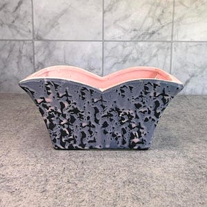 May include: A ceramic planter with a unique shape and color scheme. The exterior is a speckled gray with black accents, while the interior is a soft pink. The planter has a wide, open top and a geometric design.