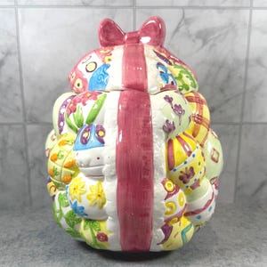 Puede incluir: Un tarro de cerámica para galletas con forma de racimo de huevos de Pascua decorados, atados con una cinta rosa brillante y un lazo. Los huevos presentan varios patrones coloridos, incluyendo flores amarillas, mariposas azules y enredaderas.