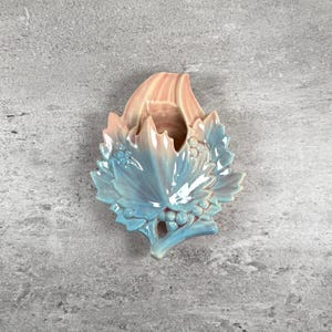 May include: A ceramic wall pocket in the shape of a leaf, with a light blue and peach color gradient. The leaf design includes detailed veins and small berry clusters, creating a decorative accent piece.