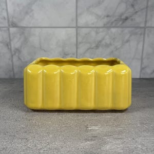 May include: A rectangular, yellow ceramic planter with a scalloped rim. The planter has vertical ribbed detailing on the sides. The container is designed for plants and adds a pop of colour to any space.